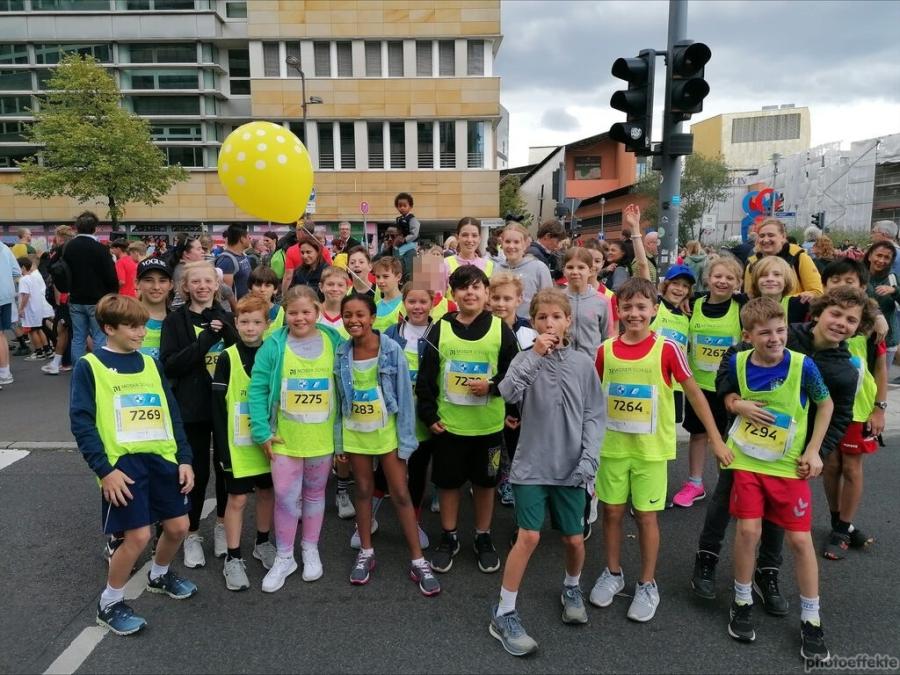 Die Kinder, die beim Marathon mitgemacht haben, stehen nebeneinander. Sie tragen Lauftrikots mit dem Logo der Schule und lächeln. Im Hintergrund sind viele Zuschauer und weitere Teilnehmer zu sehen. 