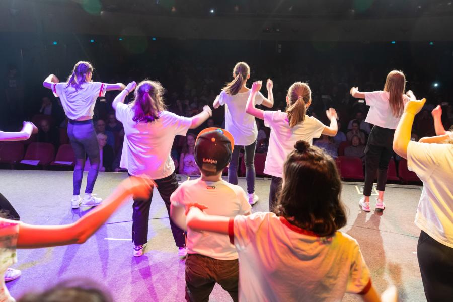 Eine Schulklasse steht im Theater auf der Bühne, im Publikum sitzen ihre Eltern. Die Kinder tragen weiße T-Shirts und führen einen Tanz auf, den sie zur Fête de Printemps einstudiert haben.  