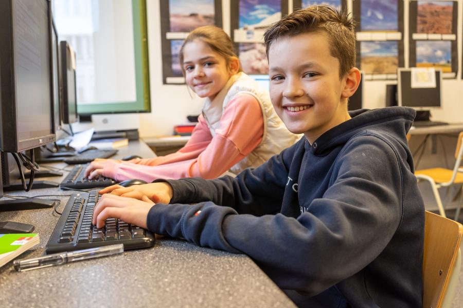 Ein Junge und ein Mädchen sitzen im Computerraum an zwei Computern. Sie lächeln und gucken in die Kamera. 