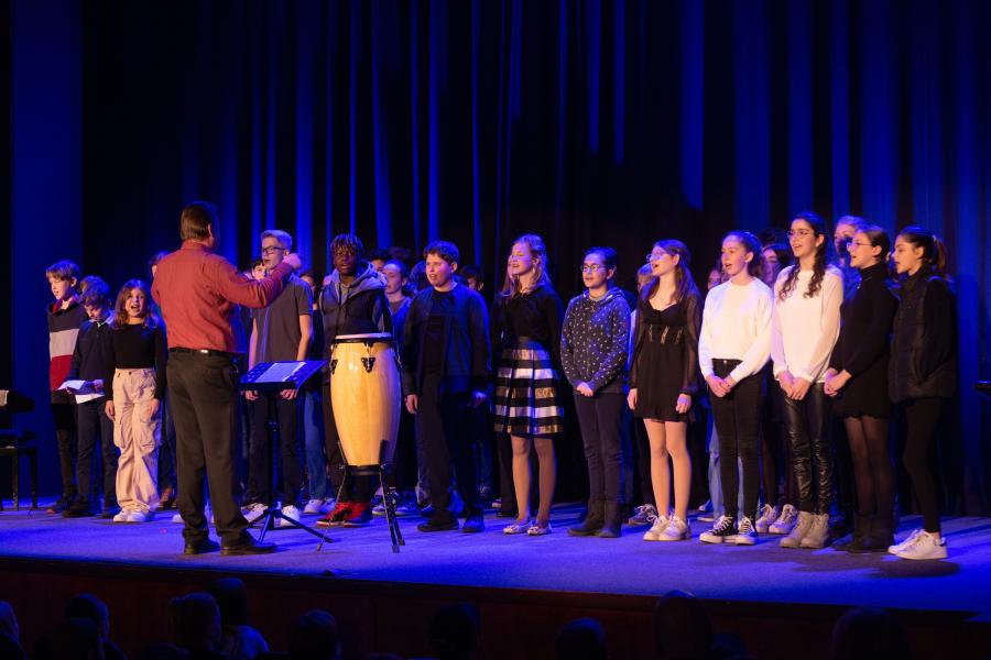 Eine Gruppe von Schülerinnen und Schülern steht auf der Bühne vom Theaterhaus. Sie singen ein einstudiertes Lied. Vor ihnen steht Herr Laas und dirigiert diesen Chor.