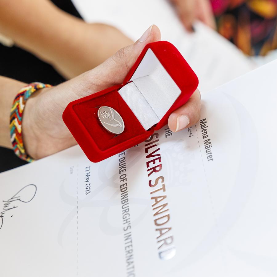 Ein Kind hält eine rote Schatulle mit der silbernen Münze des Duke of Edinburg Awards. Das Kind hält zudem eine Urkunde in der Hand.  