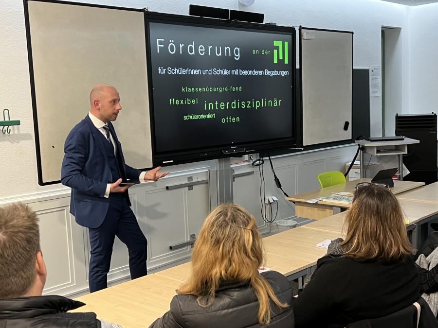 Auf dem Bild ist der vortragende Lehrer, Herr Bostanjoglo,zu sehen. Er steht vor einem digitalen Screen stellt seine Präsentation vor. Vor ihm sitzen Zuhörer und lauschen seinem Vortrag. 