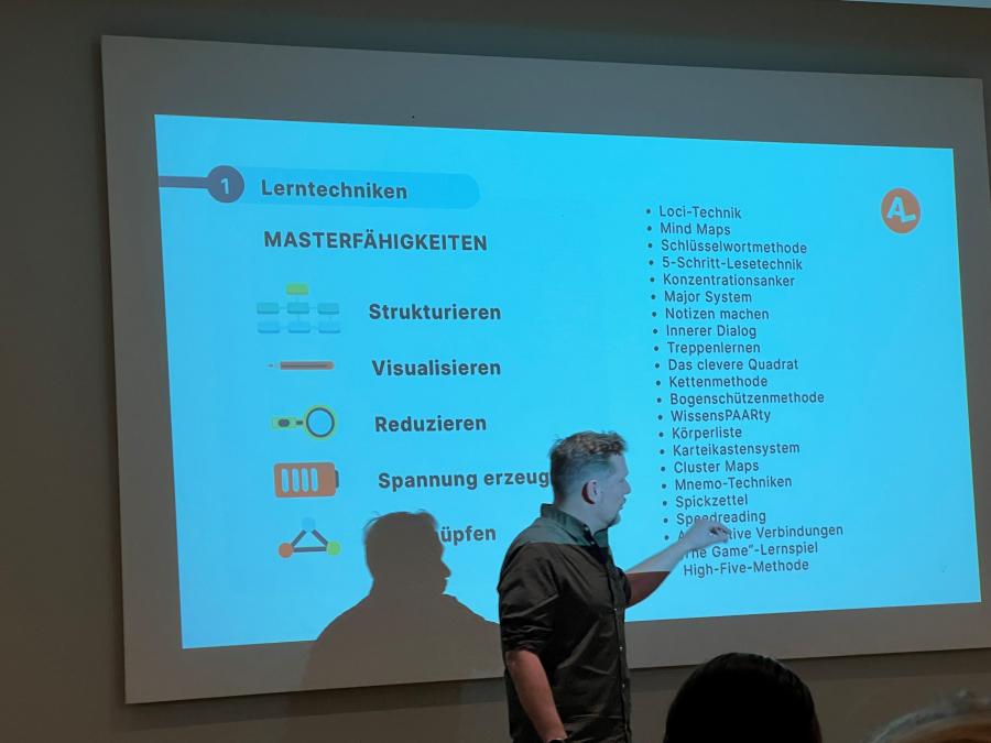 Der Referent der Lernakademie steht vor einem Bildschirm. Die zu sehende Präsentation veranschaulicht Lerntechniken.  