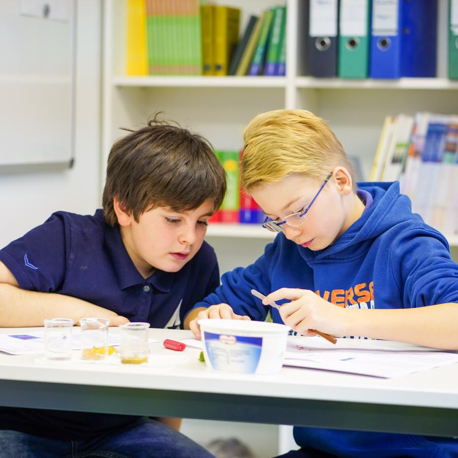 Zwei Jungen sitzen gemeinsam an einem Tisch, sie gucken gespannt auf ein Experiment, das sie gerade durchführen.  