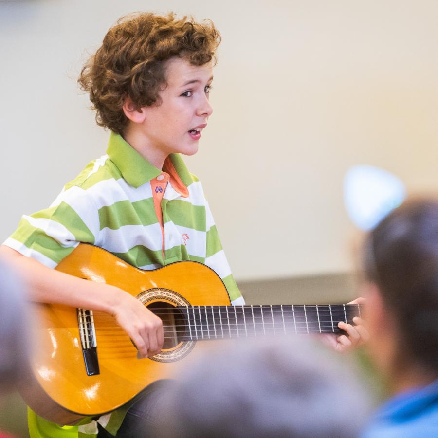 Ein kleiner Junge mit Locken und einem gestreiften T-Shirt sitzt vor einem Publikum. Er spielt auf einer Gitarre und singt dazu. 