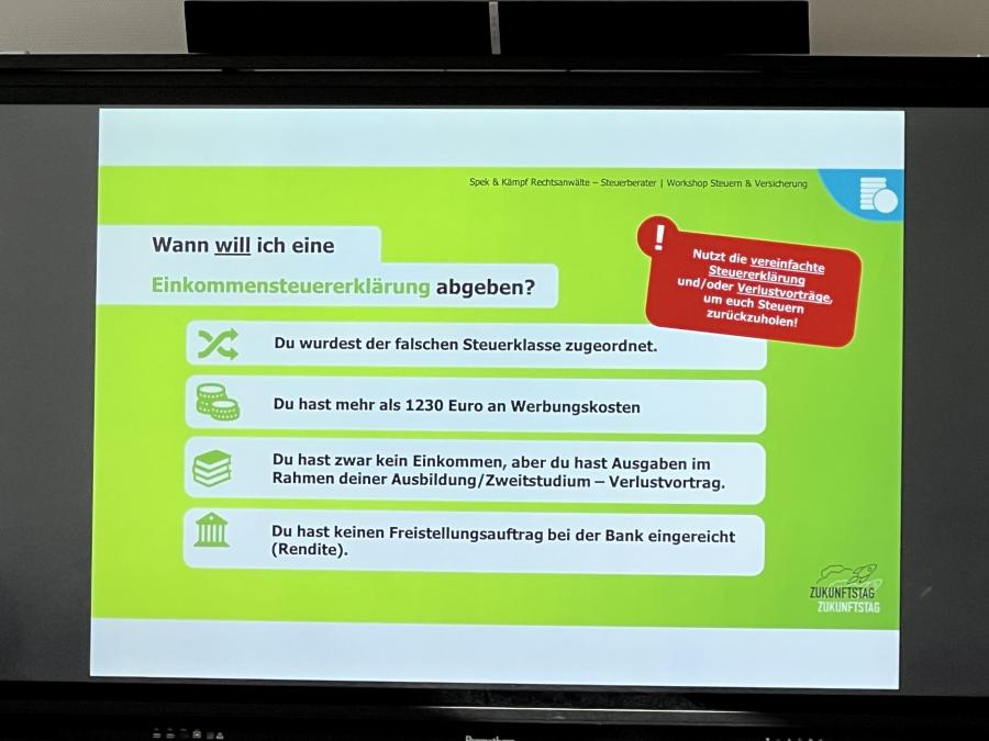 Zu sehen ist ein Bildschirm mit einer Präsentation zum Thema Steuern und Versicherungen. Der Hintergrund der Folie ist grün. Auf dem Bildschirm befinden sich Informationen zur Einkommenssteuererklärung. Die Schülerinnen und Schüler, die dem Vortrag zuhören, sind auf dem Bild nicht zu sehen.